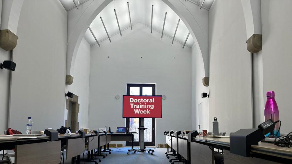 Imatge d'una sala buida amb una televisió al centre on hi ha el cartell de la Doctoral Training Week